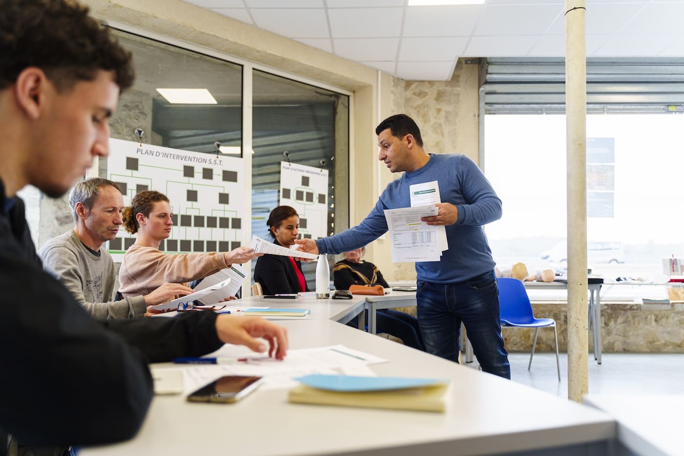 Séance de formation à l’École Gustave : un formateur BTP remet des documents à des adultes en reconversion autour d’une table de travail.