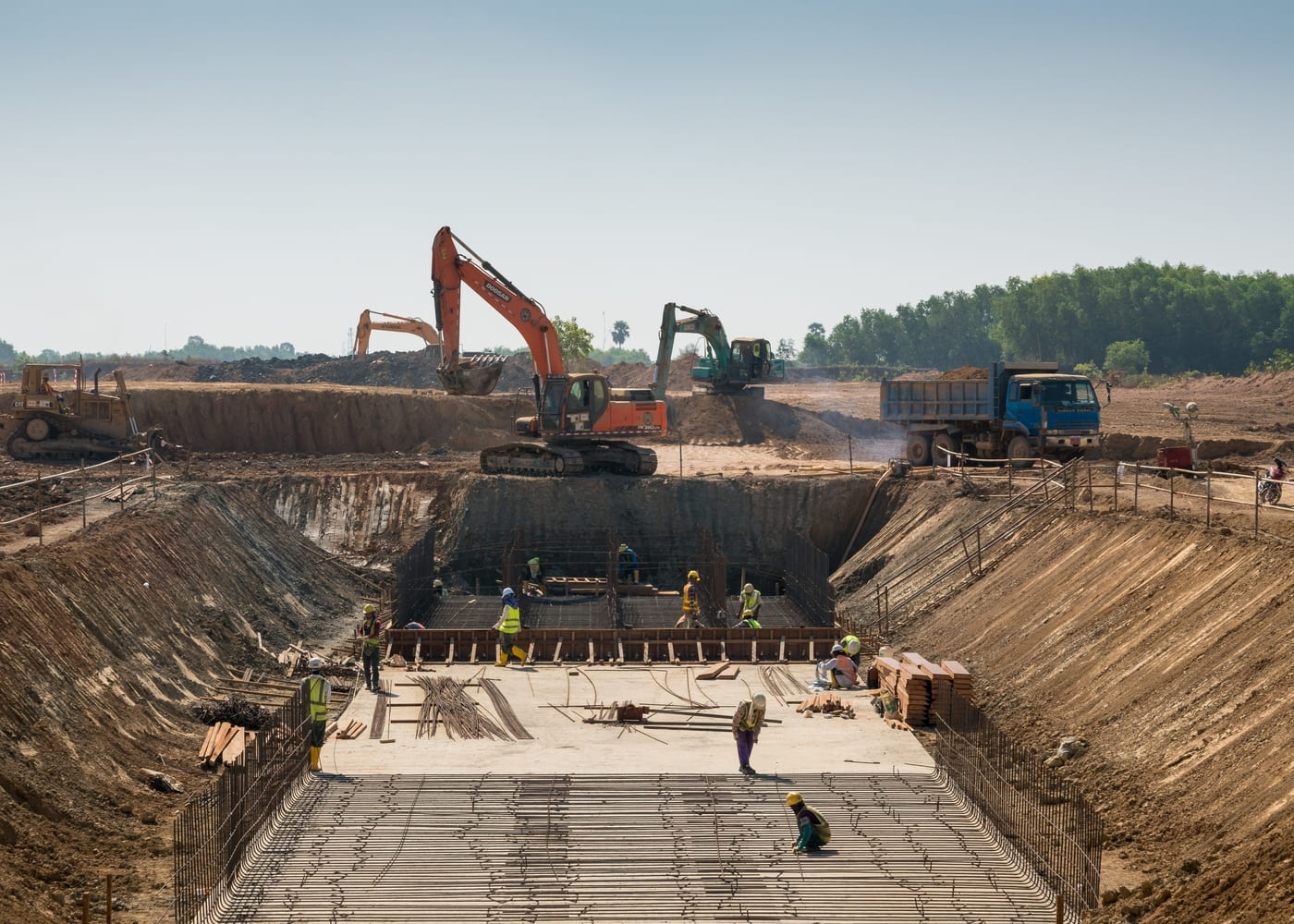 Une vue de dessus d'un grand chantier de travaux publics en cours. Des ouvriers en gilets de sécurité et casques travaillent dans une tranchée profonde sur des structures en acier et des coffrages. Des pelleteuses et un camion-benne sont postés sur le terrain supérieur.