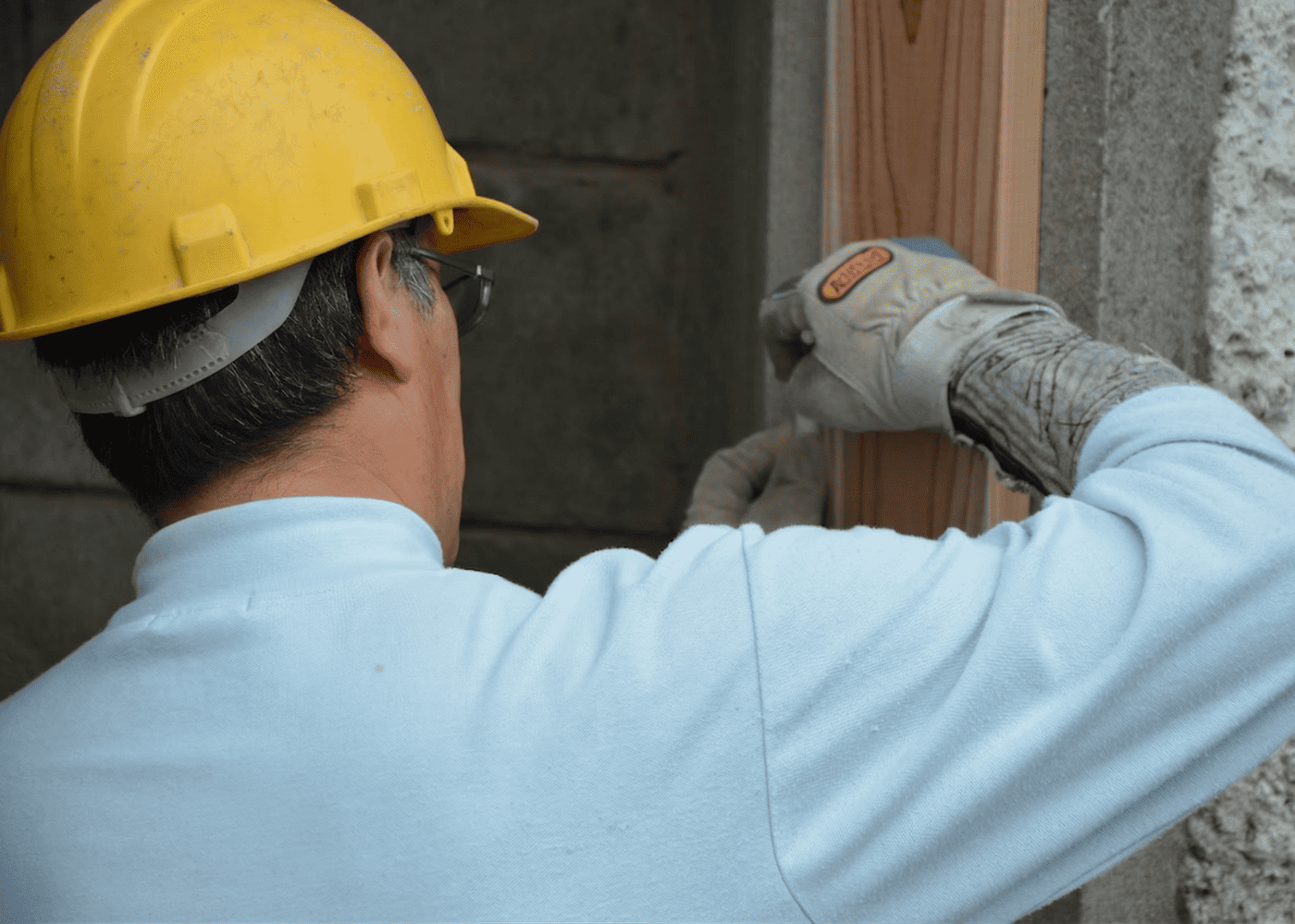 Menuisier installateur de dos portant un casque jaune et des gants, en train de poser ou régler un élément de menuiserie sur un chantier.
