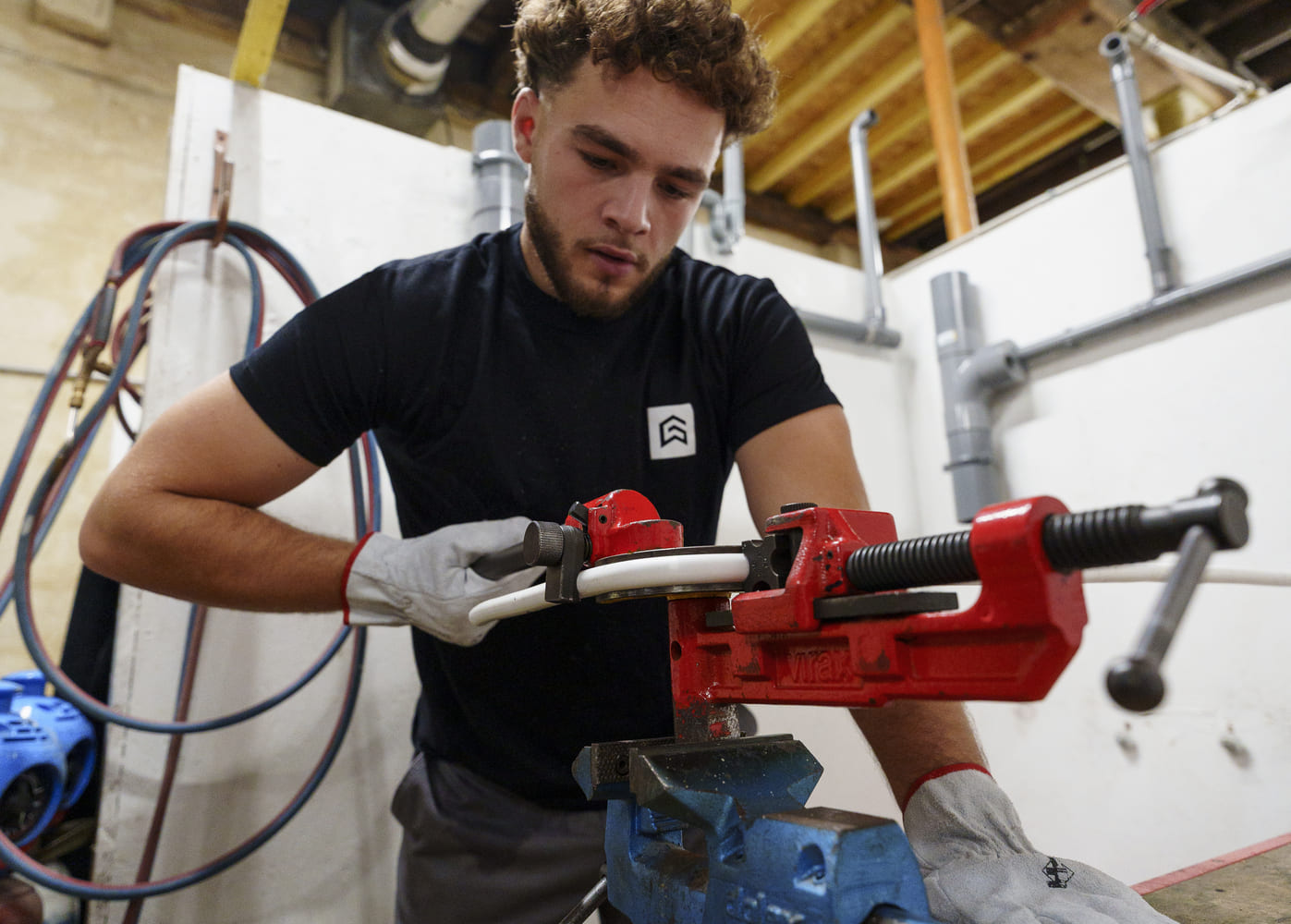 Jeune apprenti aux cheveux bouclés en t-shirt noir logo gris, gants de travail gris, utilise une pince à sertir rouge sur un tube blanc à l’étau bleu dans un atelier plomberie, avec tuyaux et outils en fond.