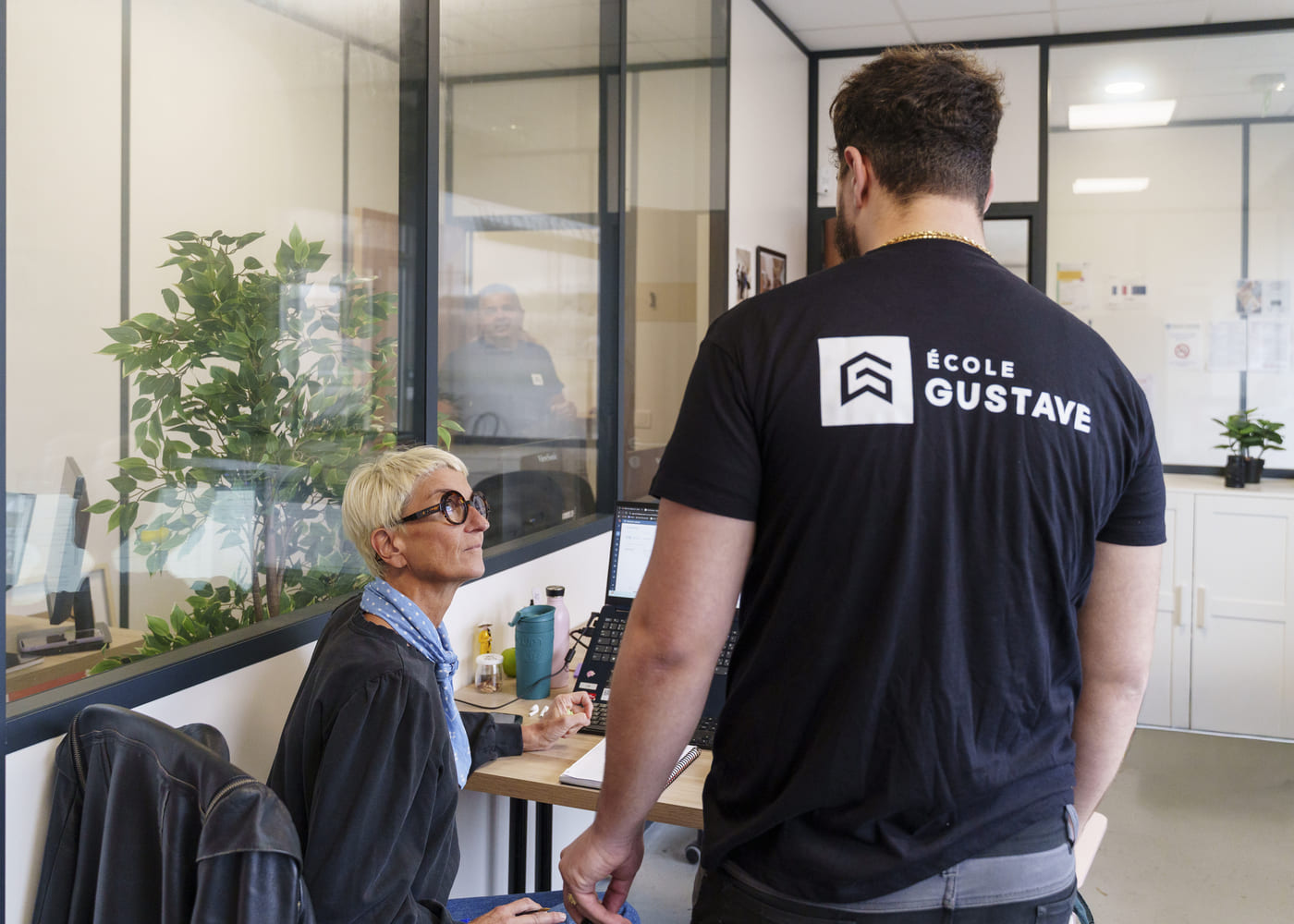 Une formatrice ou conseillère pédagogique de l'École Gustave, assise à son poste de travail dans un bureau moderne et lumineux, discute avec un apprenant (homme de dos, portant le t-shirt siglé "ÉCOLE GUSTAVE"). La scène illustre un moment d'échange, d'orientation ou de suivi de parcours. Sur le bureau en bois clair se trouvent un ordinateur portable ouvert, une gourde, un thermos et une plante verte en pot. Des cloisons vitrées définissent l'espace de bureau, derrière lesquelles un autre homme est visible assis. Un blouson en cuir noir est posé sur la chaise de la femme.