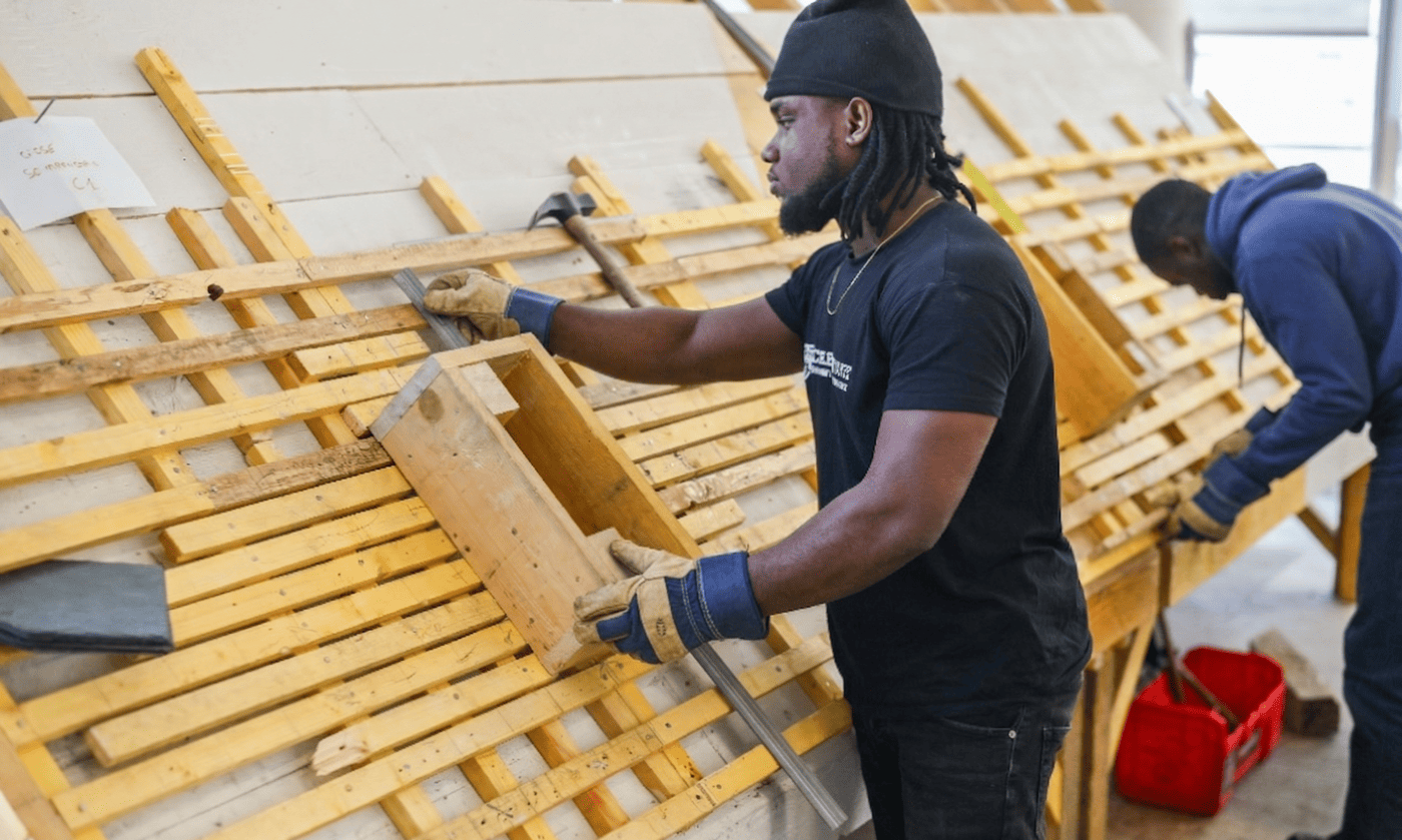 Un étudiant équipé de gants de protection, portant un bonnet et un t-shirt noir avec le logo de l'École Gustave, prend des mesures avec une équerre métallique sur une structure d'apprentissage de toiture en bois. En arrière-plan, un autre apprenti en tenue de travail bleue s'exerce sur la même installation.