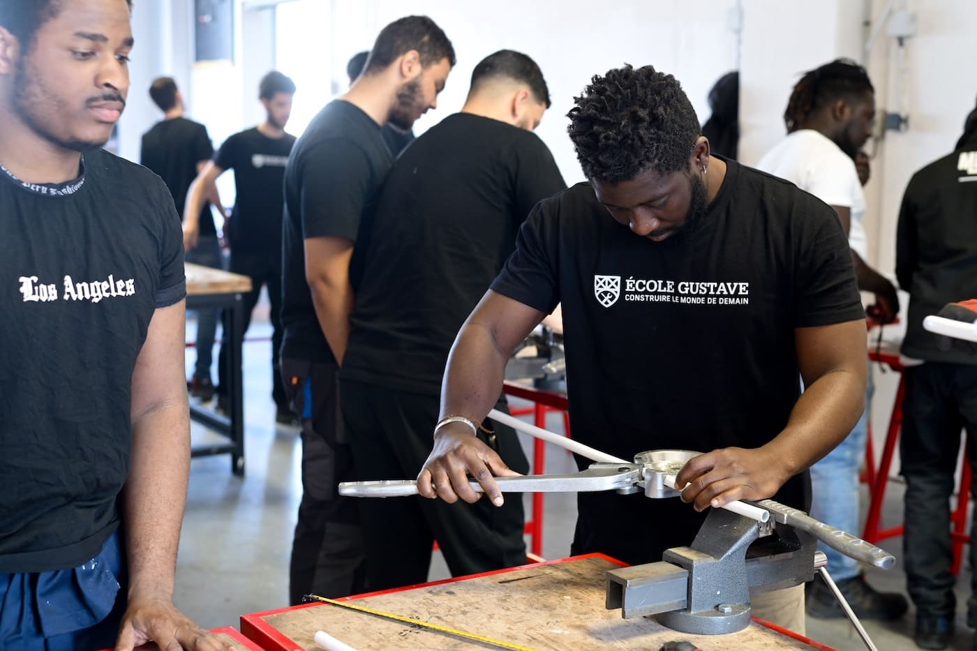 Un jeune homme noir, portant un t-shirt noir avec le logo blanc visible "ÉCOLE GUSTAVE" et le slogan "CONSTRUIRE LE MONDE DE DEMAIN", utilise une cintreuse de tubes manuelle sur un tuyau blanc fixé dans un étau sur un établi en bois. Il est concentré sur son geste technique. D'autres étudiants en formation sont visibles en arrière-plan, flous.