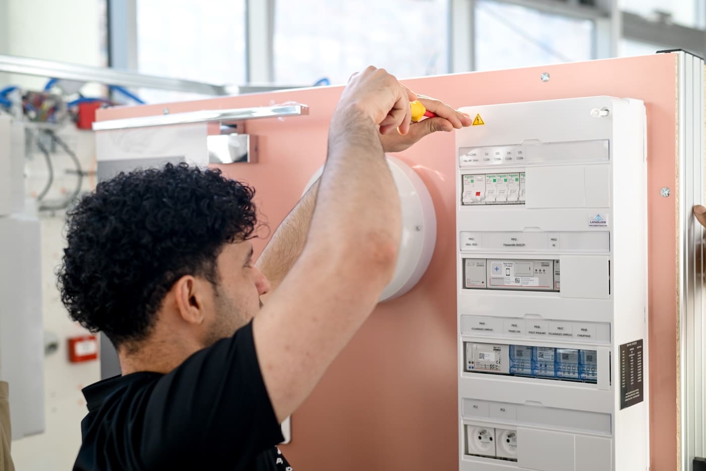 Un apprenti électricien en t-shirt noir utilise un tournevis isolé pour ajuster les disjoncteurs d'un tableau électrique blanc, fixé sur une cloison rose lors d'un exercice pratique à l'École Gustave.