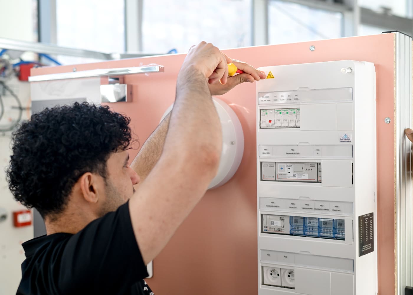Un apprenant de l'École Gustave en t-shirt noir utilise un tournevis d'électricien isolé pour intervenir sur les disjoncteurs d'un tableau électrique pédagogique.