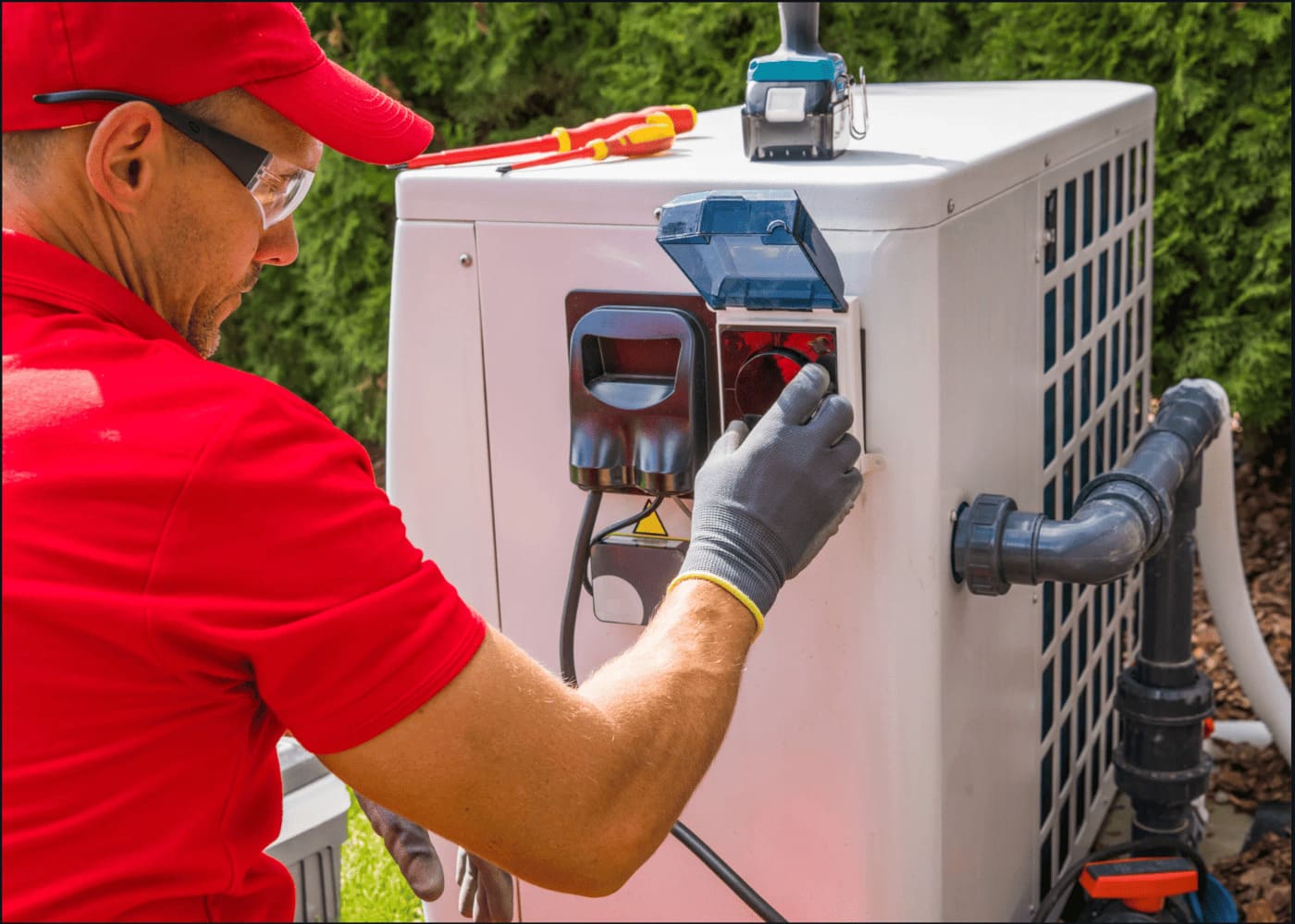 Un technicien de maintenance CVC en polo rouge, portant des lunettes de sécurité et des gants, ajuste un réglage sur le panneau de commande latéral d'une unité extérieure de pompe à chaleur air-eau. Des tournevis et une perceuse sont posés sur le dessus de l'appareil blanc dans un jardin ensoleillé.