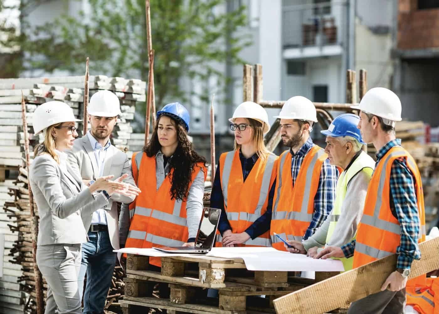 Une femme cheffe d’équipe en costume donne des instructions à un groupe d’ouvriers et d’ouvrières du BTP sur un chantier, autour de plans et d’un ordinateur portable posés sur une palette.
