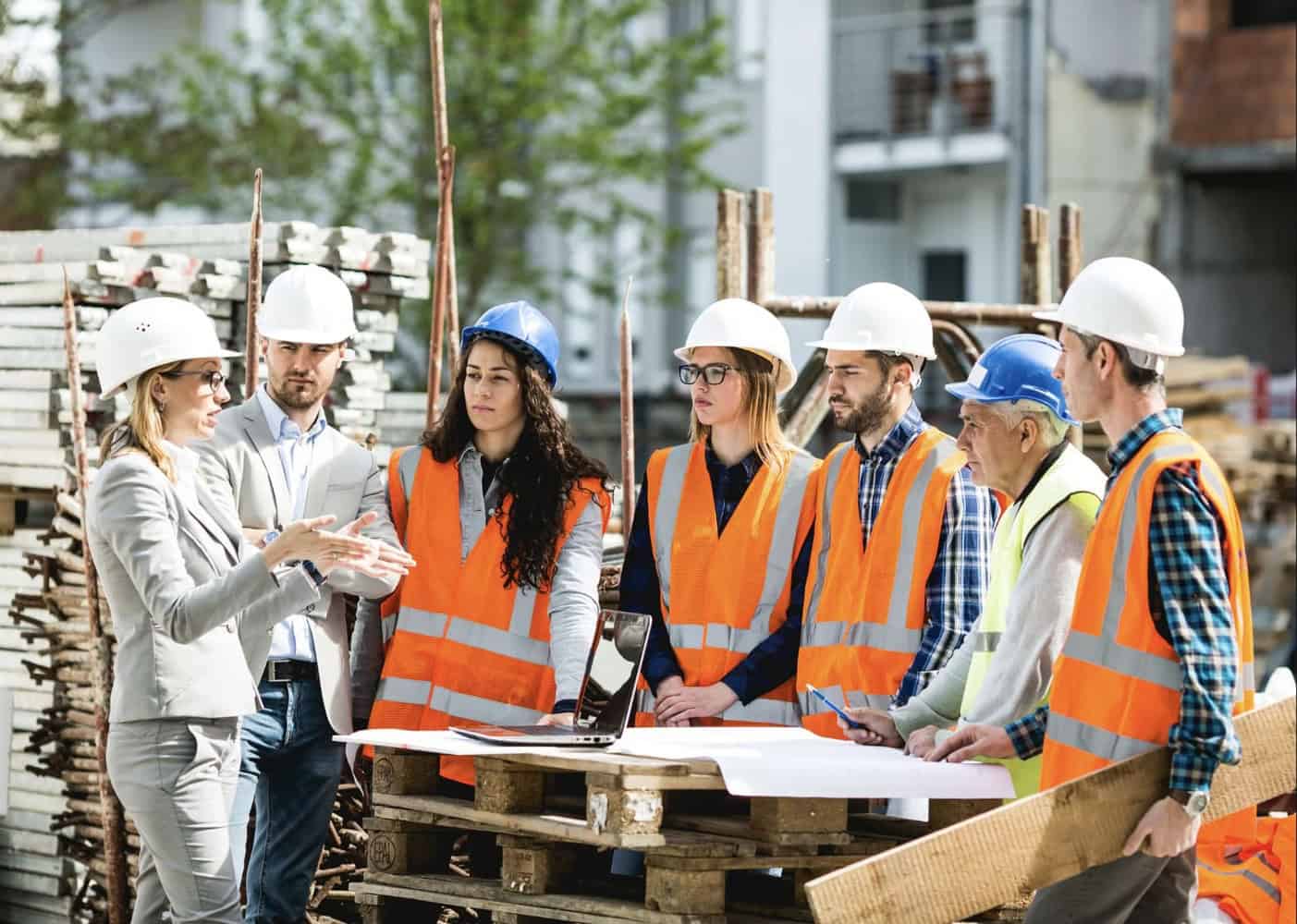 Une femme cheffe d’équipe en costume donne des instructions à un groupe d’ouvriers et d’ouvrières du BTP sur un chantier, autour de plans et d’un ordinateur portable posés sur une palette.
