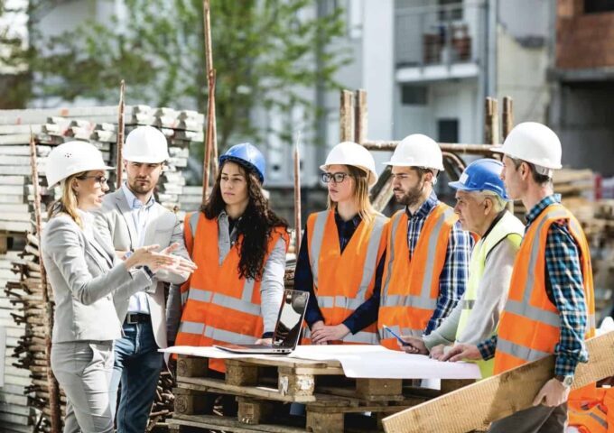 Une femme cheffe d’équipe en costume donne des instructions à un groupe d’ouvriers et d’ouvrières du BTP sur un chantier, autour de plans et d’un ordinateur portable posés sur une palette.