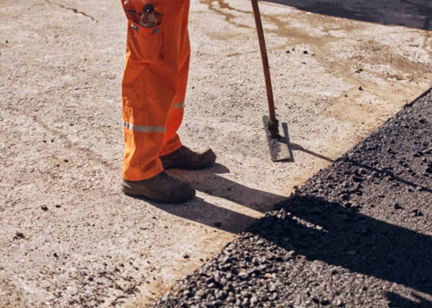 Maçon VRD équipé d'un pantalon haute visibilité orange et de chaussures de sécurité, maniant un outil de nivellement sur un chantier d'aménagement de voirie goudronnée.