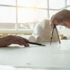 Mains traçant un plan d'architecte avec un compas et un stylo, casque de chantier blanc posé sur la table — reconversion professionnelle dans le BTP