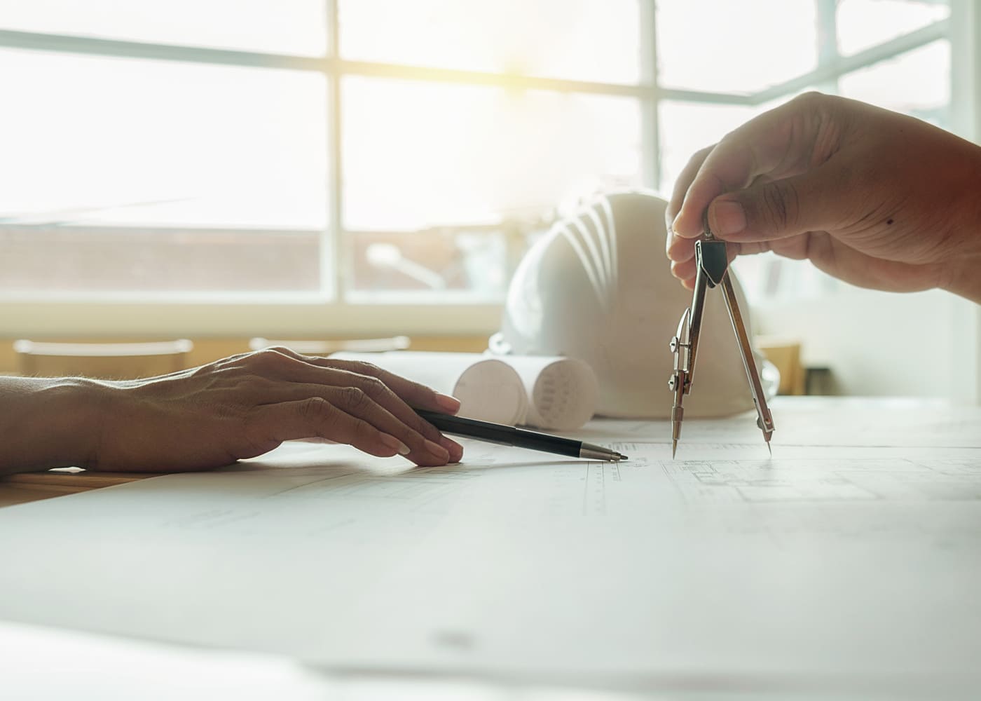 Mains traçant un plan d'architecte avec un compas et un stylo, casque de chantier blanc posé sur la table — reconversion professionnelle dans le BTP