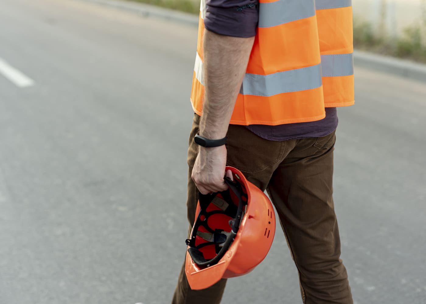 Un ouvrier du BTP porte un gilet orange à bandes réfléchissantes. Il est de dos et tient un casque de chantier orange dans sa main droite. Il se trouve sur une route asphaltée.