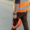 Un maçon VRD de dos portant un gilet de sécurité orange et tenant un casque de chantier à la main sur une route goudronnée.
