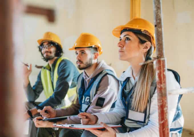 Trois apprenants équipés de casques de chantier suivent une formation pratique en intérieur avec carnet de notes et matériel de travail.