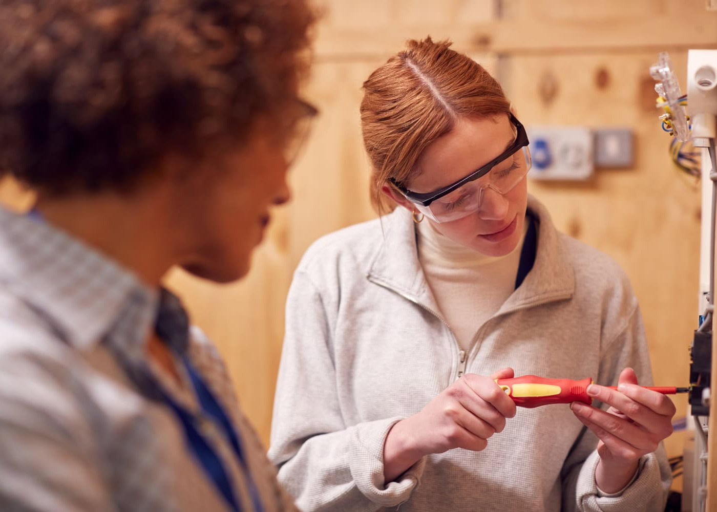 Une jeune femme portant des lunettes de protection manipule un tournevis isolé sur une installation électrique, observée par sa formatrice.