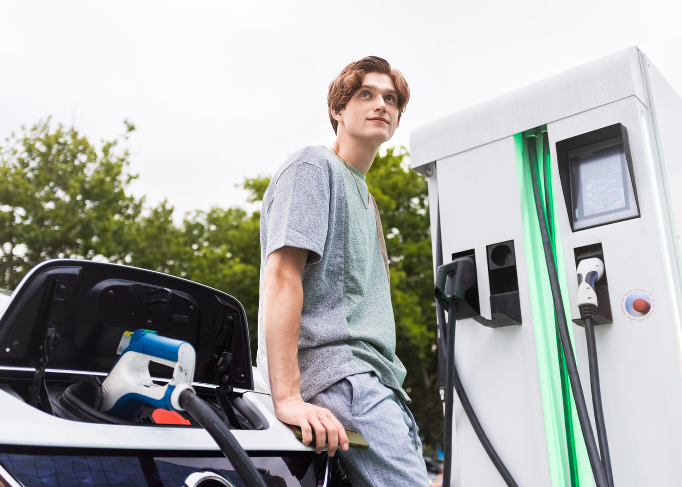 Un jeune homme appuyé contre sa voiture électrique pendant la recharge sur une borne publique rapide IRVE.