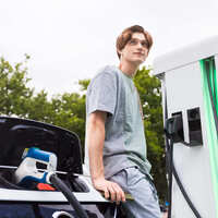 Un jeune homme appuyé contre sa voiture électrique pendant la recharge sur une borne publique rapide IRVE.